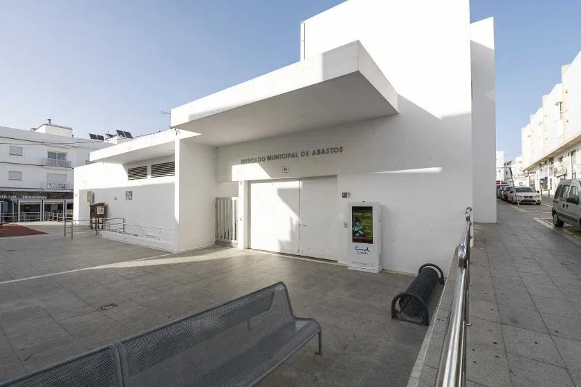 A white public building opens onto a paved plaza with benches and a covered entrance.