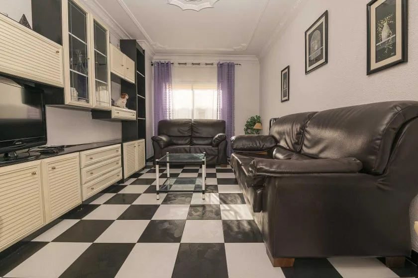 A living room with dark seating, a glass coffee table, and a black-and-white checkered floor.