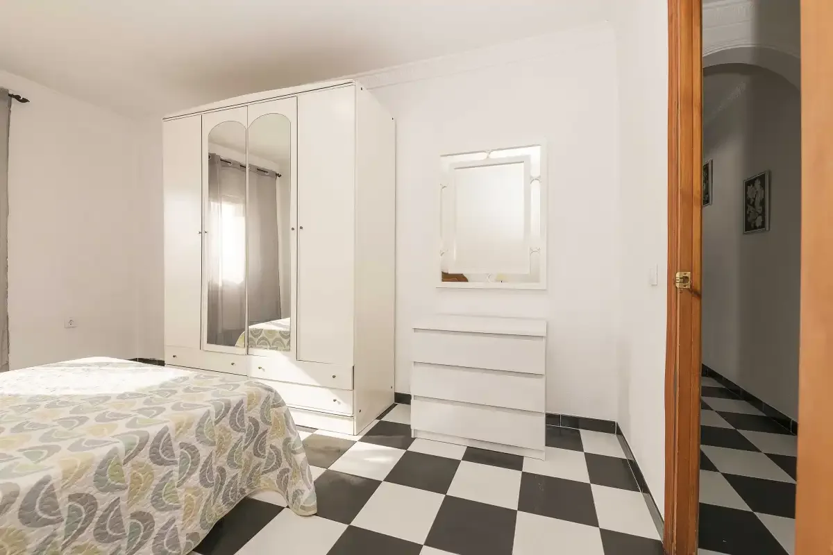 A bedroom with a bed, mirrored wardrobe, and dresser on a checkerboard floor.