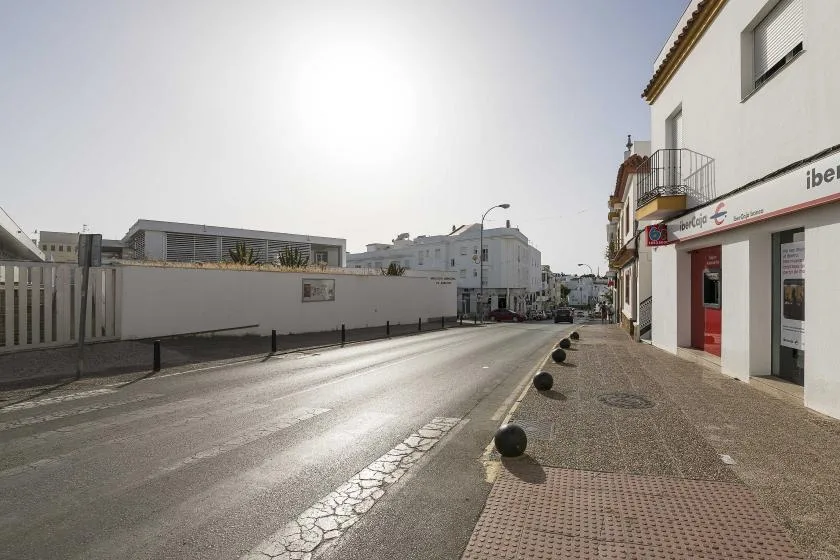 A sunlit street runs past a white building and a paved sidewalk.