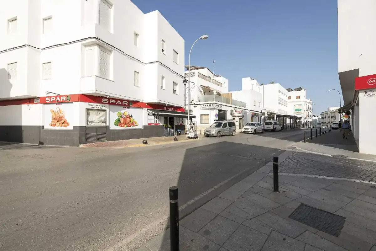 A sunny urban street lined with white buildings, shops, and parked vehicles.