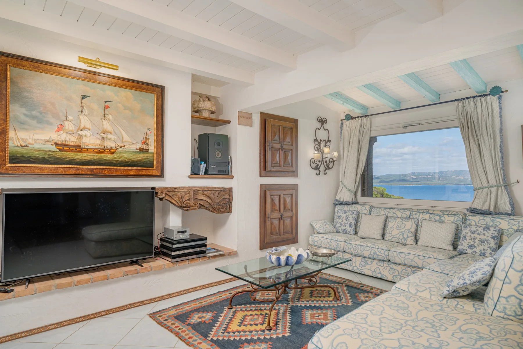 A living room with patterned seating, a glass coffee table, and a large window overlooking the water.