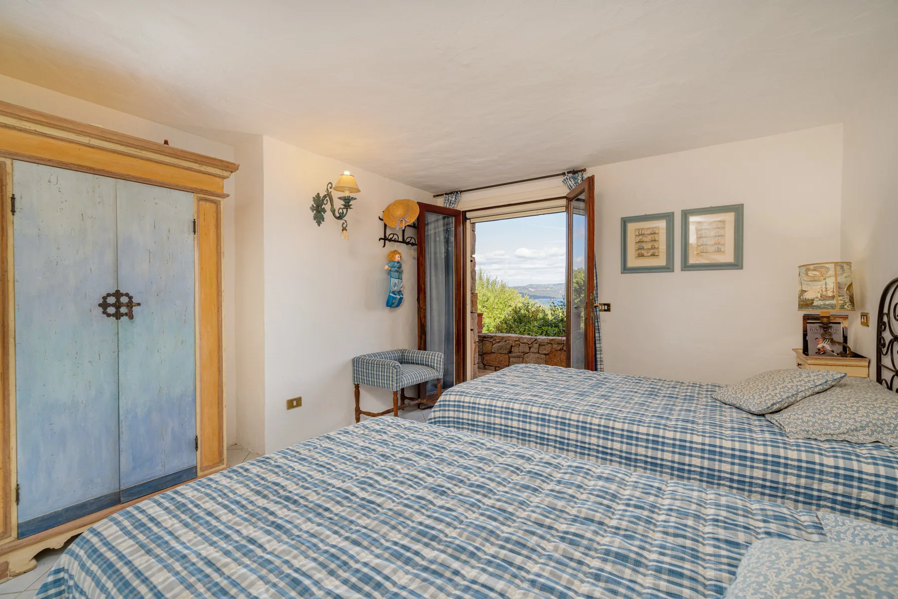 A bedroom with beds beside an open window overlooking greenery and distant hills.