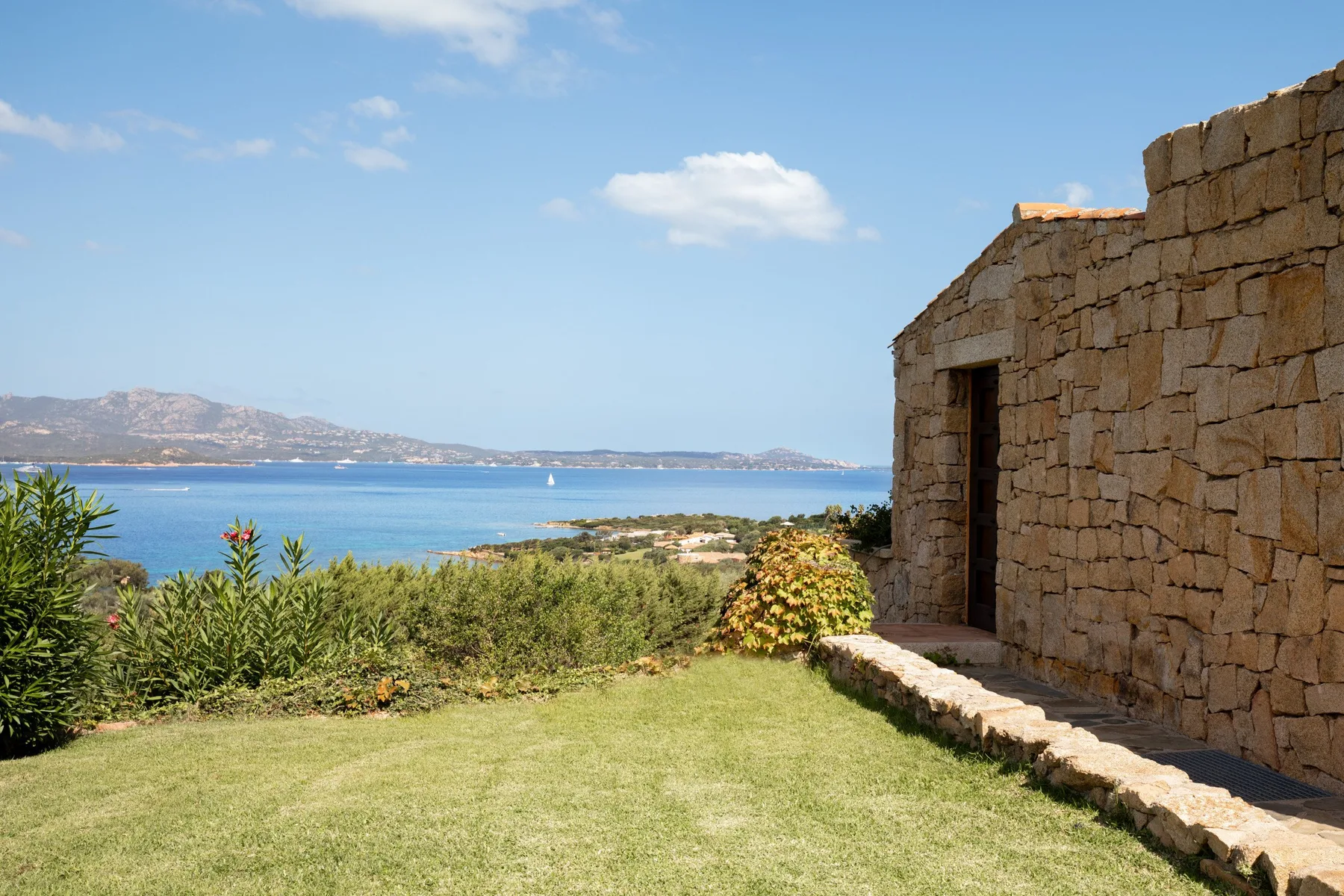 A stone building beside a lawn overlooks the sea and distant hills.