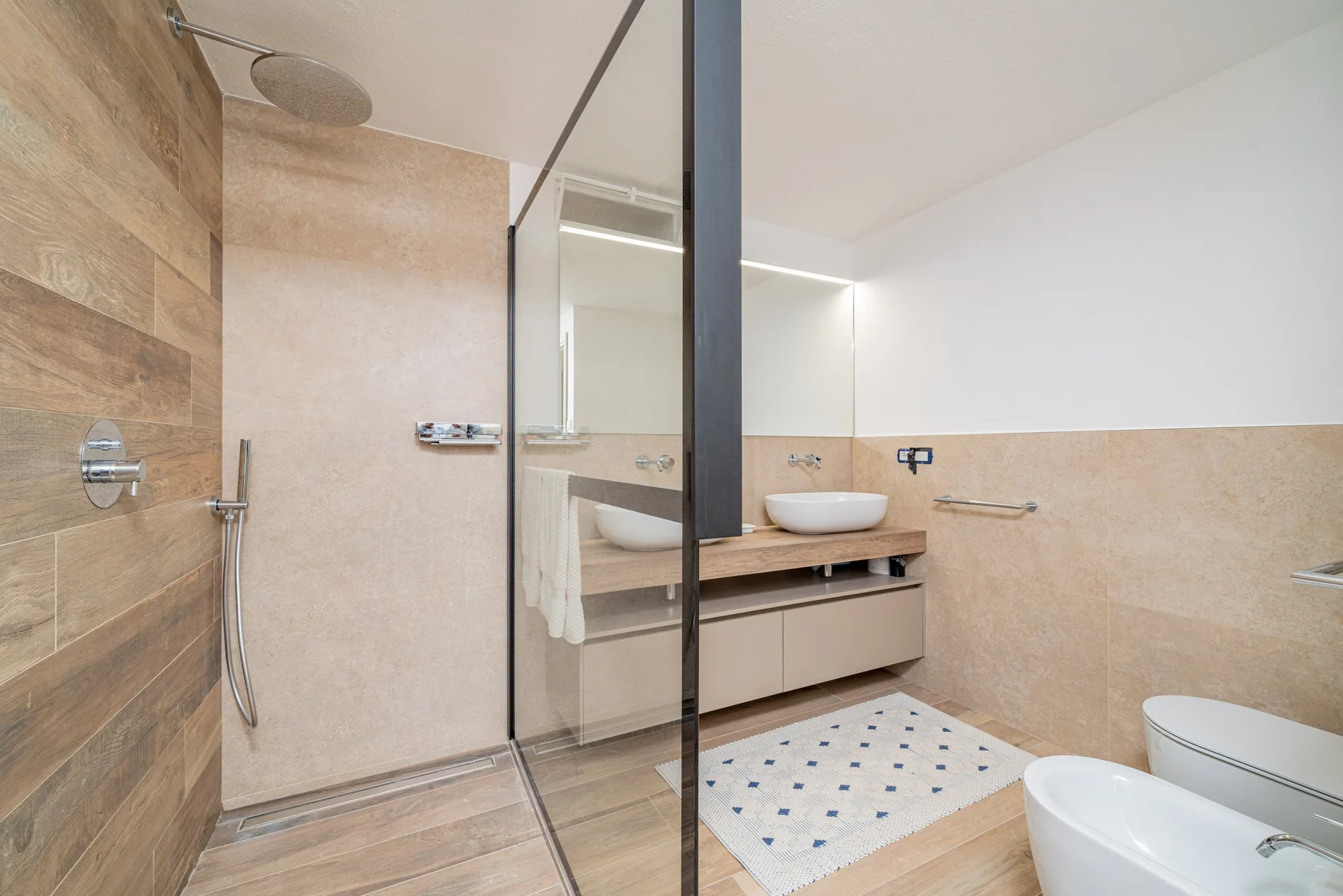A modern bathroom with a glass shower, vanity, and patterned floor mat.