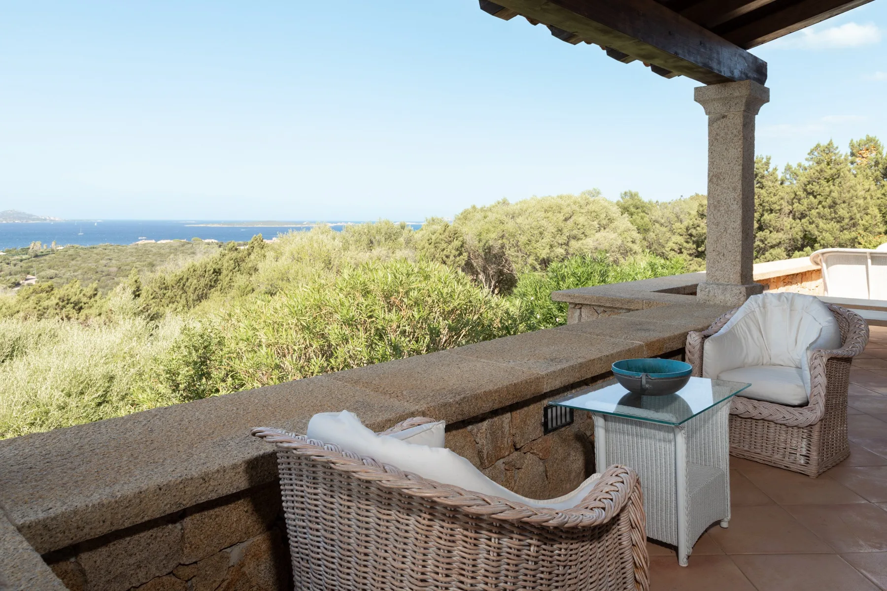 A covered terrace with wicker seating overlooks green shrubs and the sea.