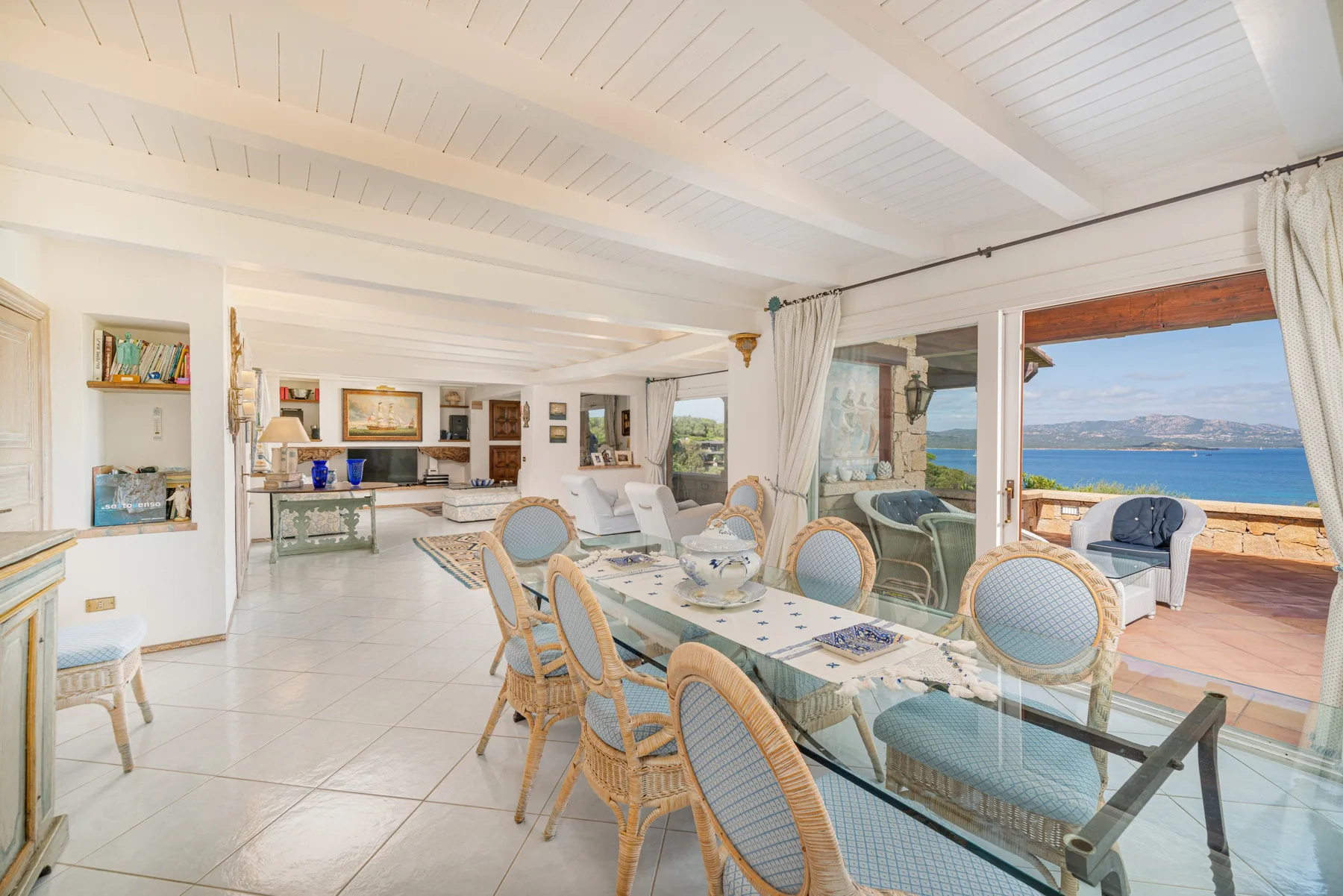 An open dining and living area has a glass table beside large windows overlooking the sea.