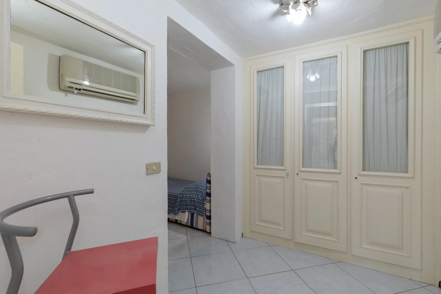 A room with pale tiled flooring, tall paneled doors, and a bed visible through an opening.
