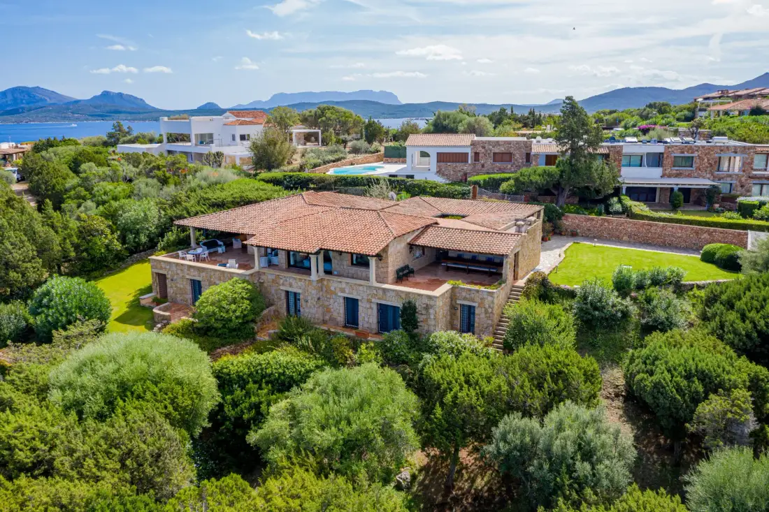 A large house sits amid dense greenery, with water and mountains in the distance.