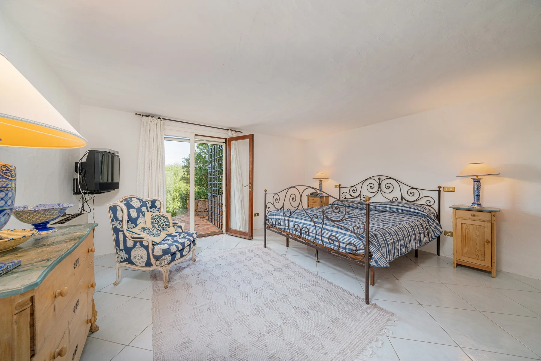 A bright bedroom with an iron bed, an upholstered armchair, and an open balcony door.