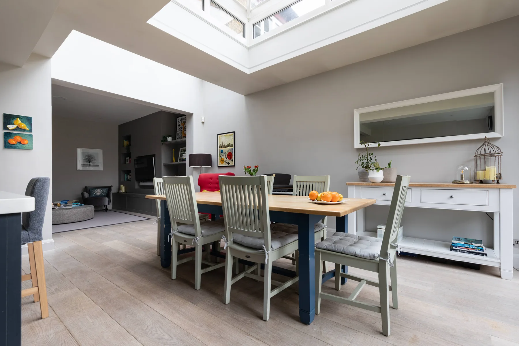 A dining area with a long table and chairs sits beneath a large skylight.