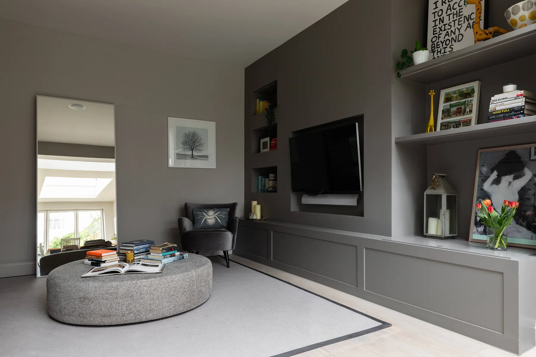 A living room with a wall-mounted television, built-in shelving, and a round ottoman.