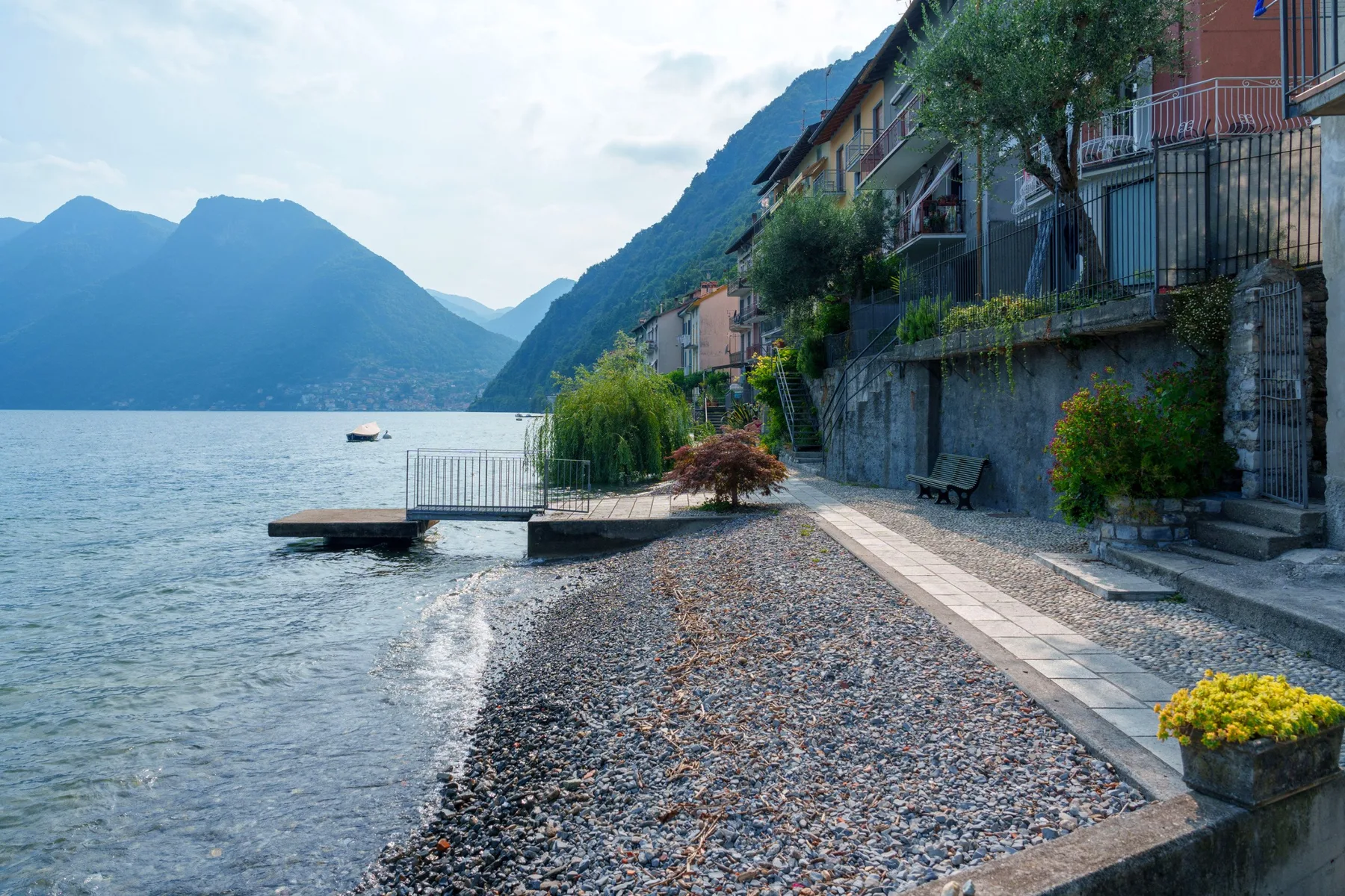 A lakeside pebble shore with a walkway beside buildings and mountain views.