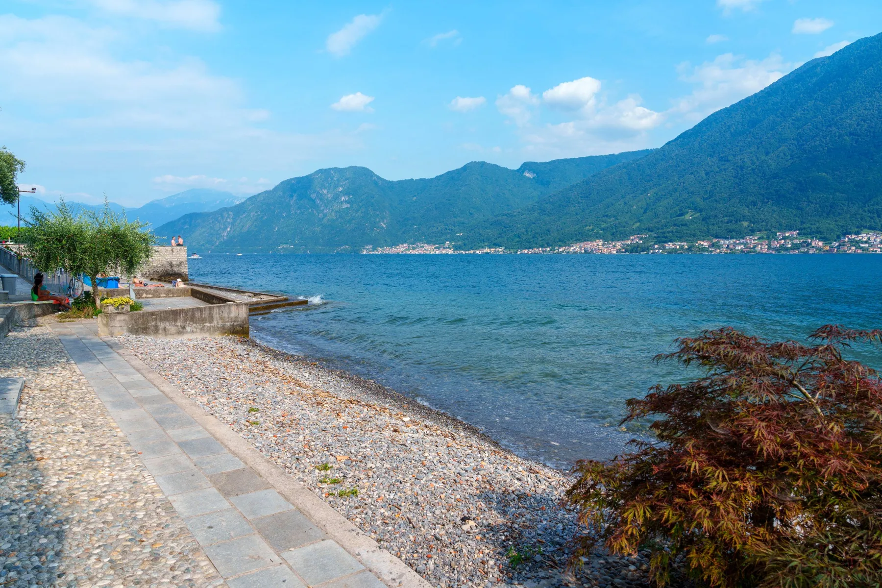 A lakeside shore with a paved path, pebbles, and mountains across the water.