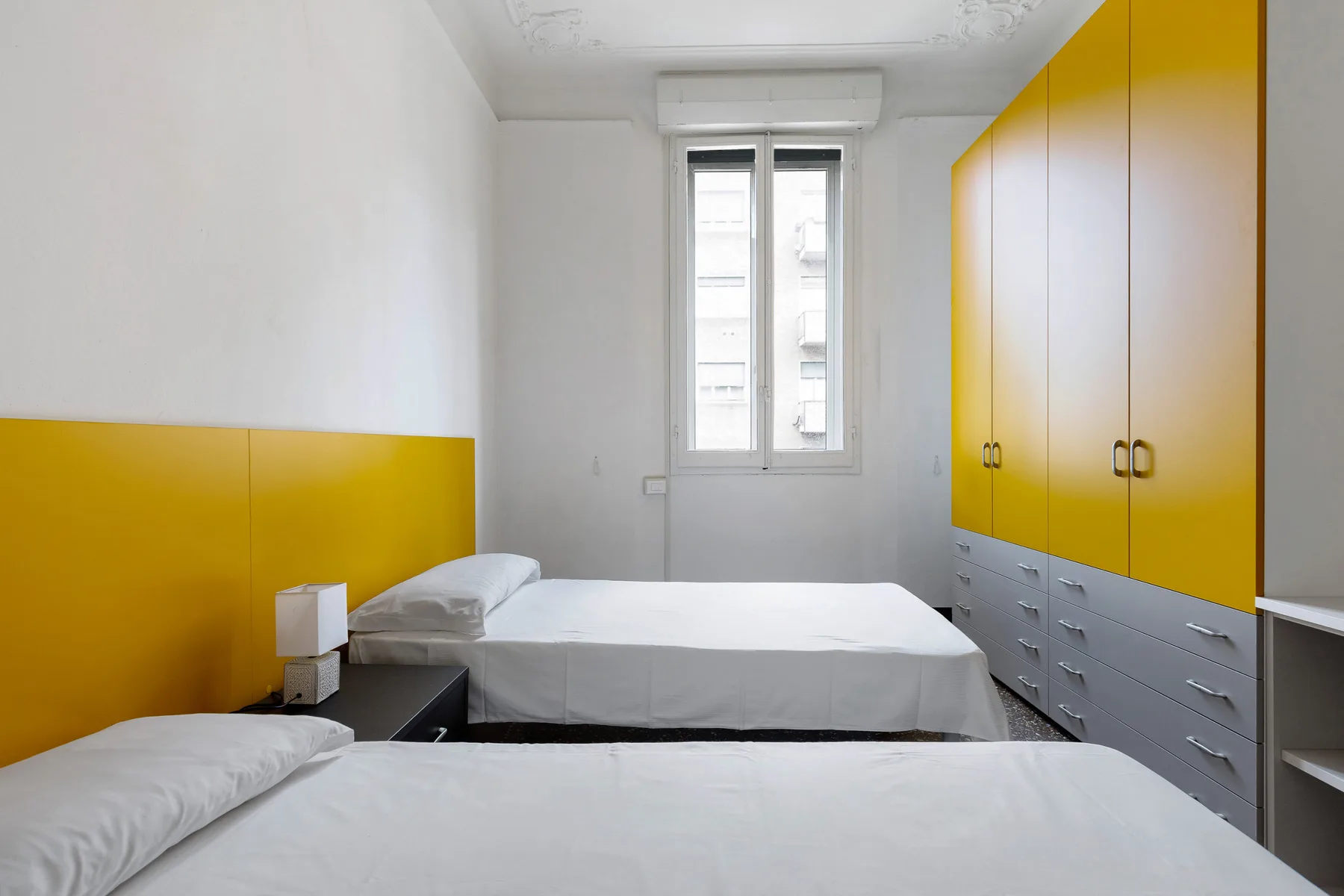 A bedroom with two beds, a central window, and yellow-and-gray built-in storage.