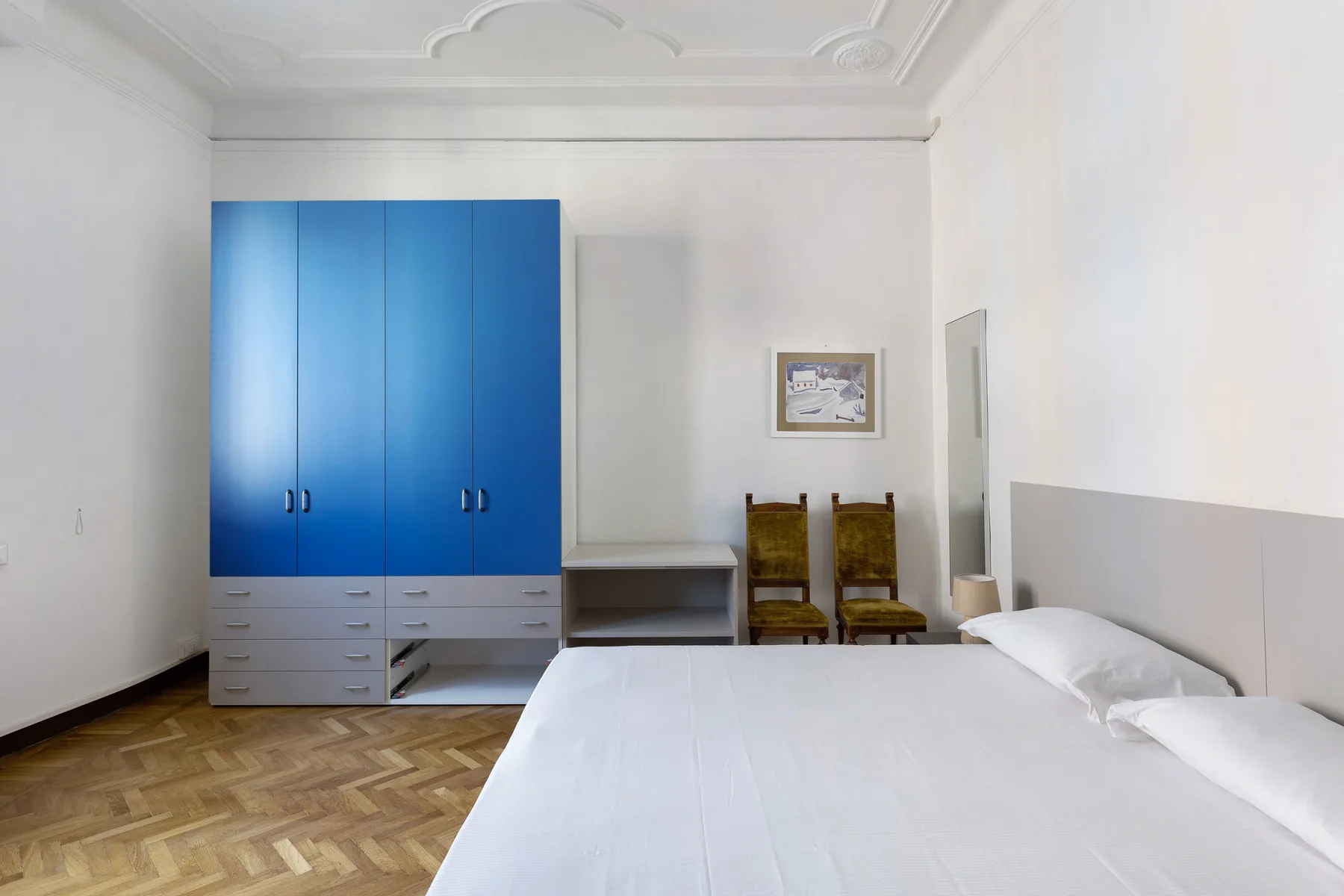 A bedroom with a bed, blue wardrobe, and two chairs.