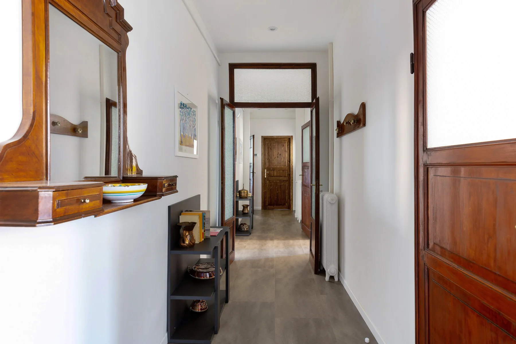 A narrow hallway with white walls, wooden doors, and a small shelving unit.