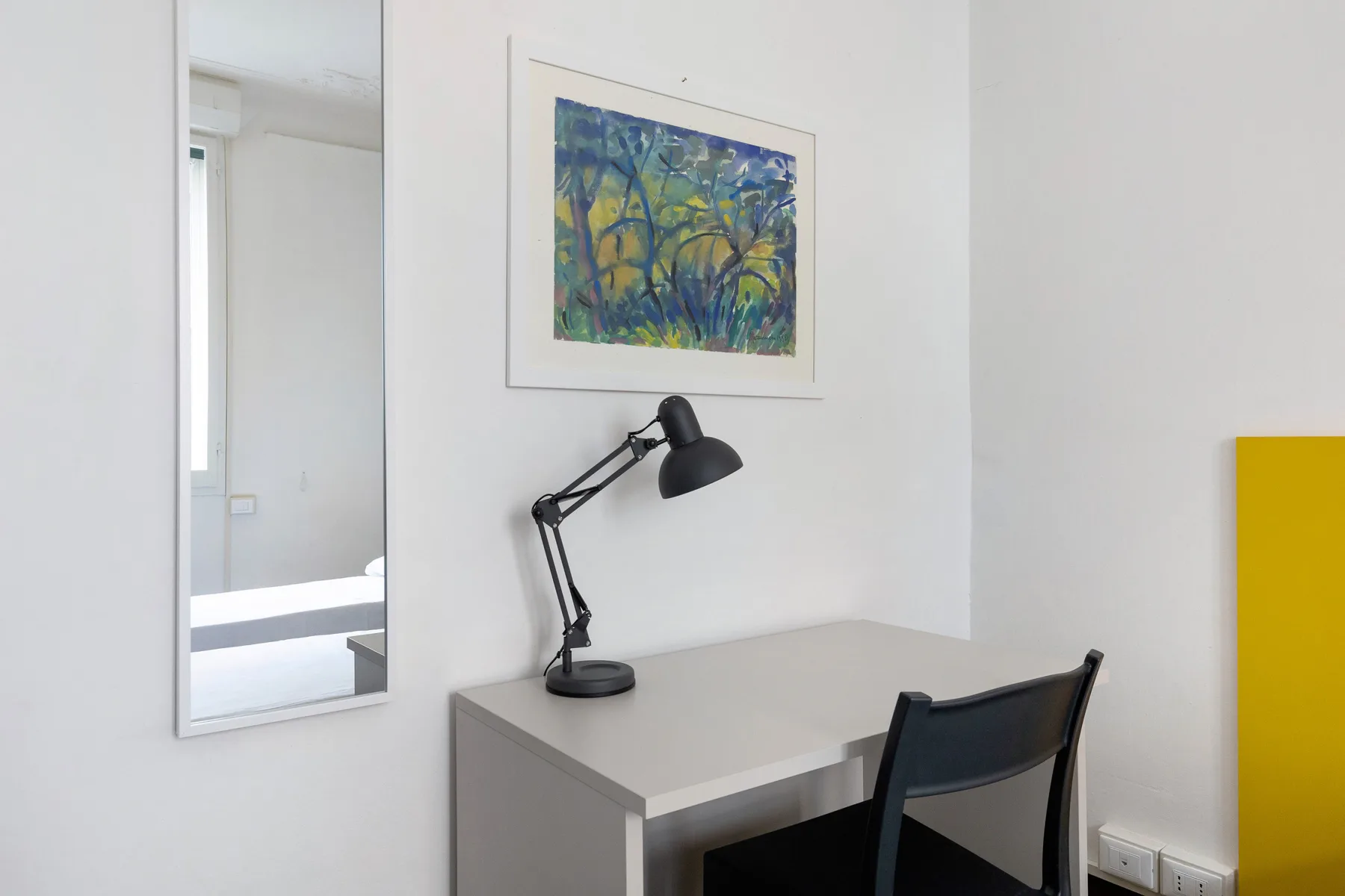A desk and chair sit beneath a framed painting in a bright room.