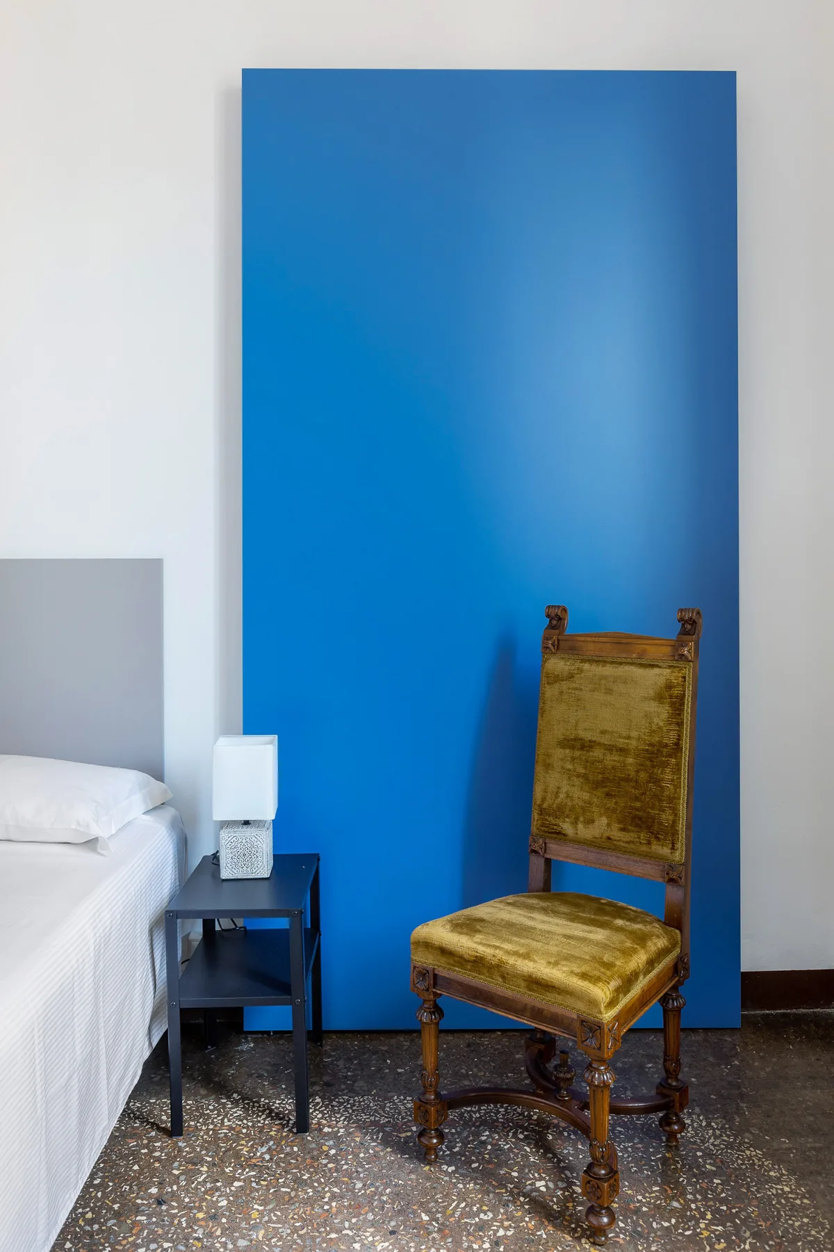 A bedroom with a bed, a small bedside table, and a wooden chair against a blue wall.