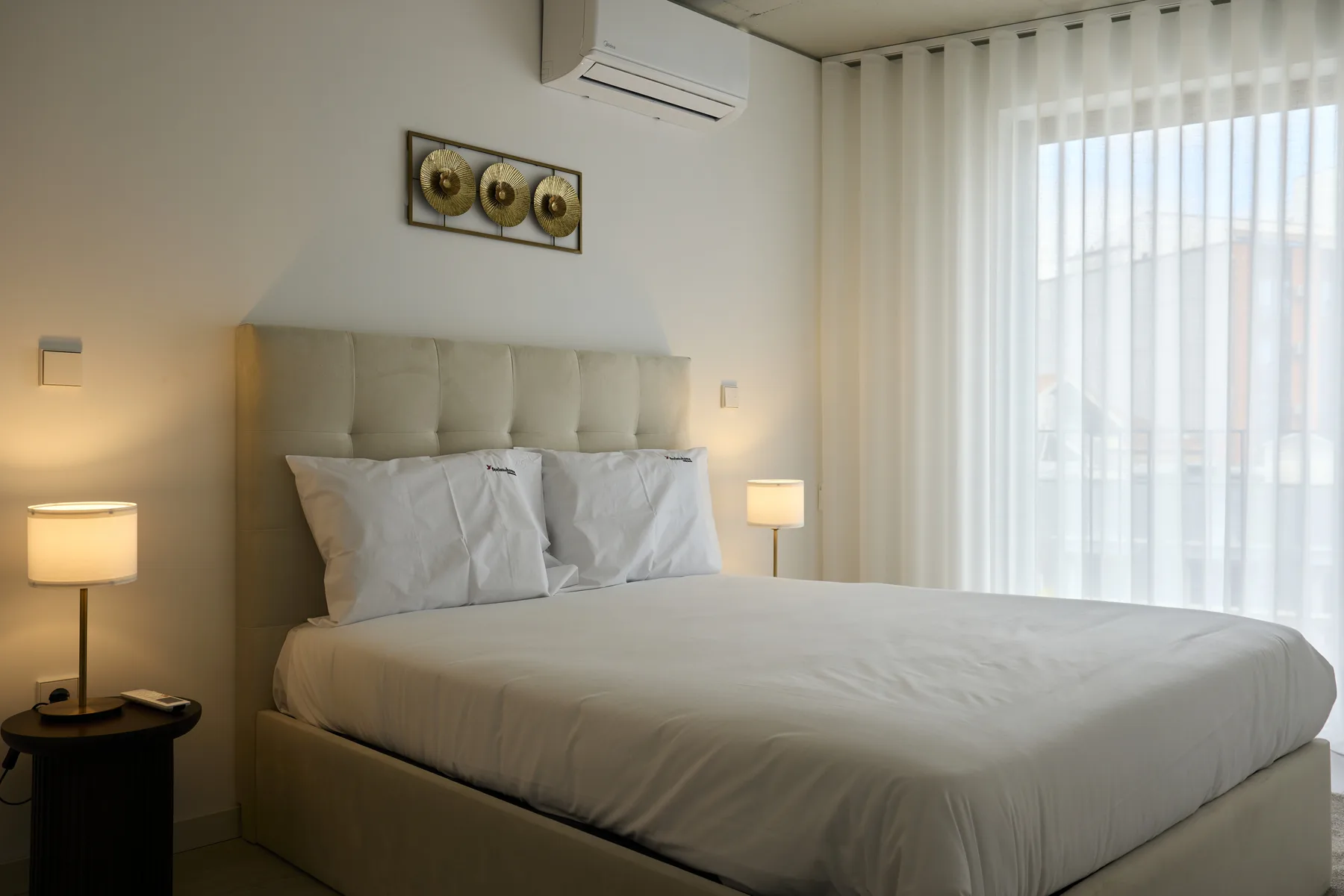A bedroom with a large bed beside a bright window with sheer curtains.