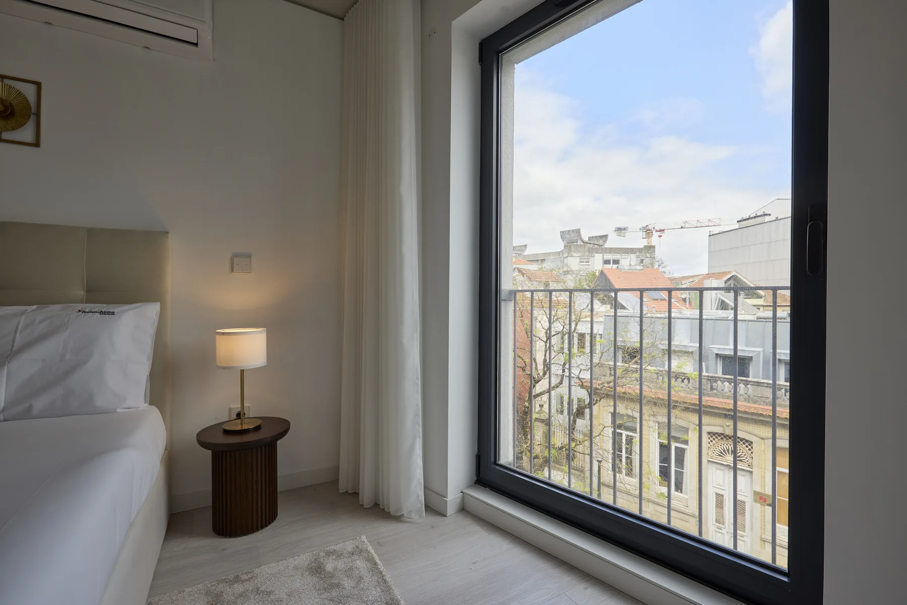 A bedroom with a bed, bedside table, and large window overlooking nearby buildings.