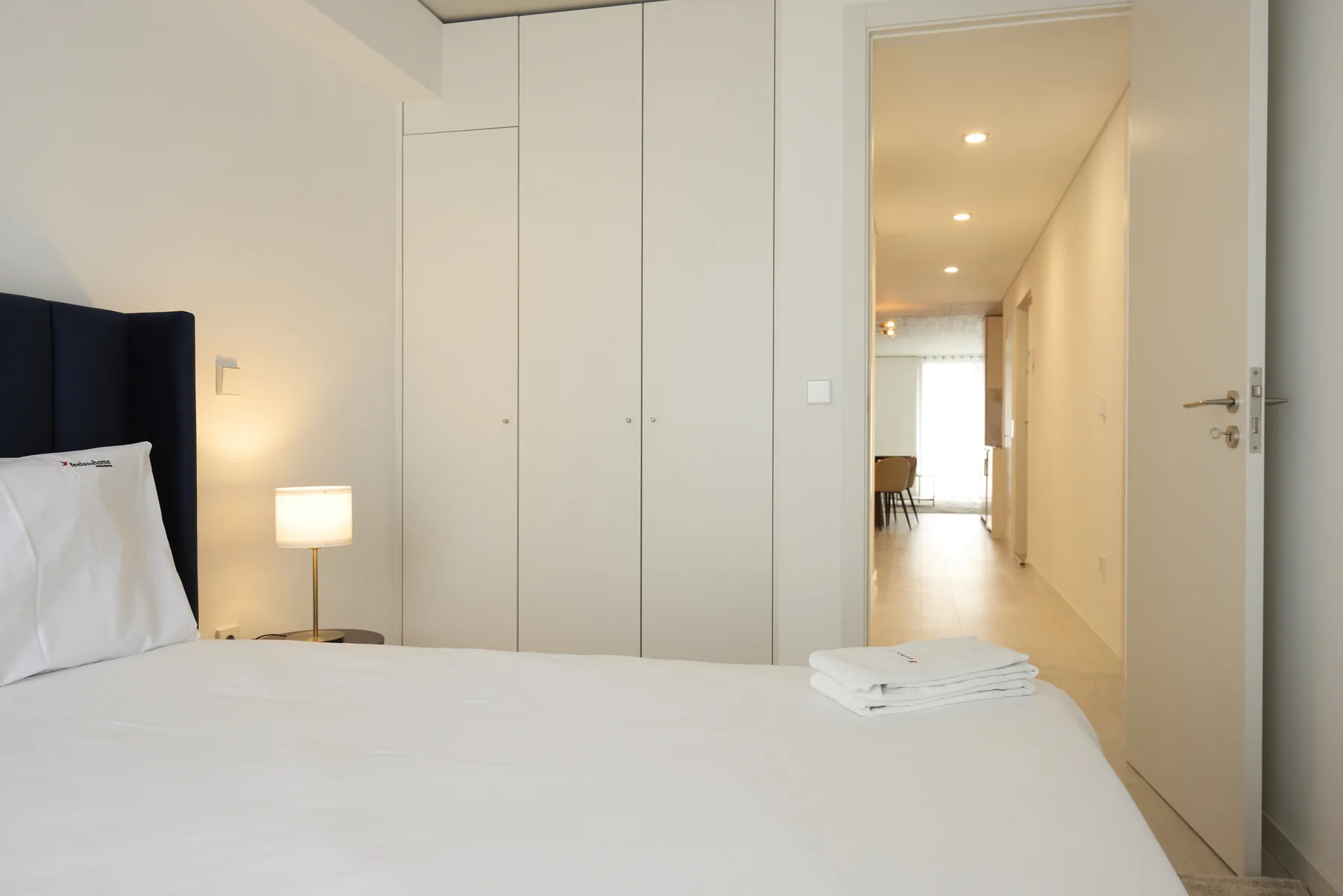 A bright bedroom with a neatly made bed and an open doorway to a hallway.