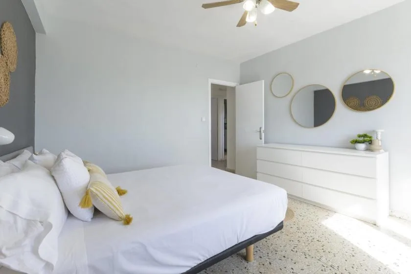 A bright bedroom with white bedding and a white dresser beneath round mirrors.