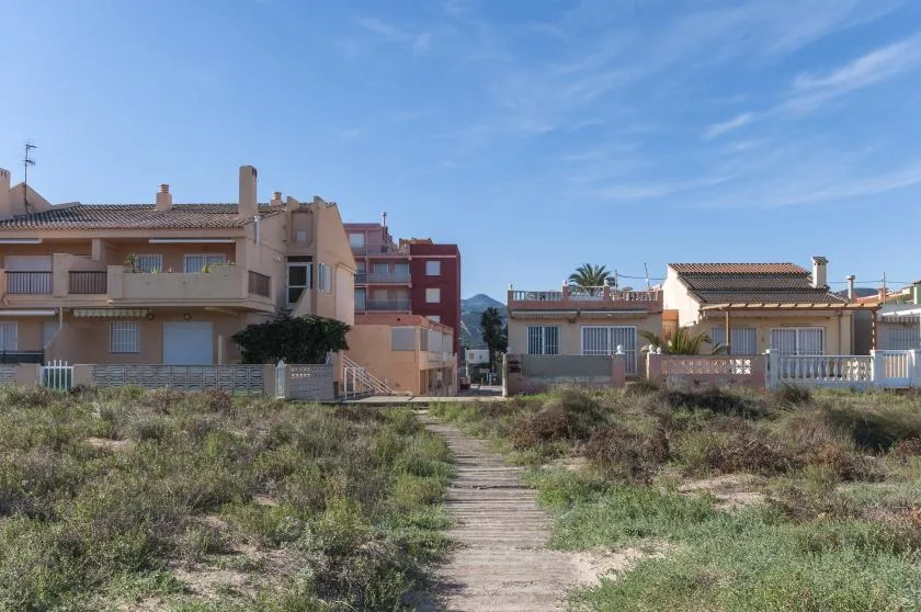 A dirt path crosses scrubby ground toward a row of low-rise buildings.