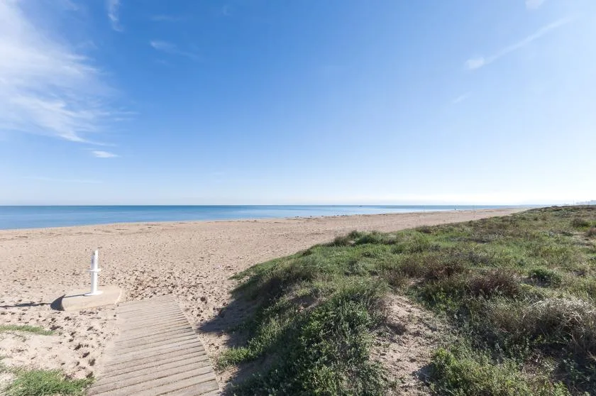 A sandy beach meets the sea beside a grassy dune and wooden boardwalk.