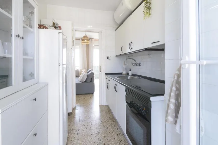 A narrow white kitchen with dark countertops and a view into the living area.