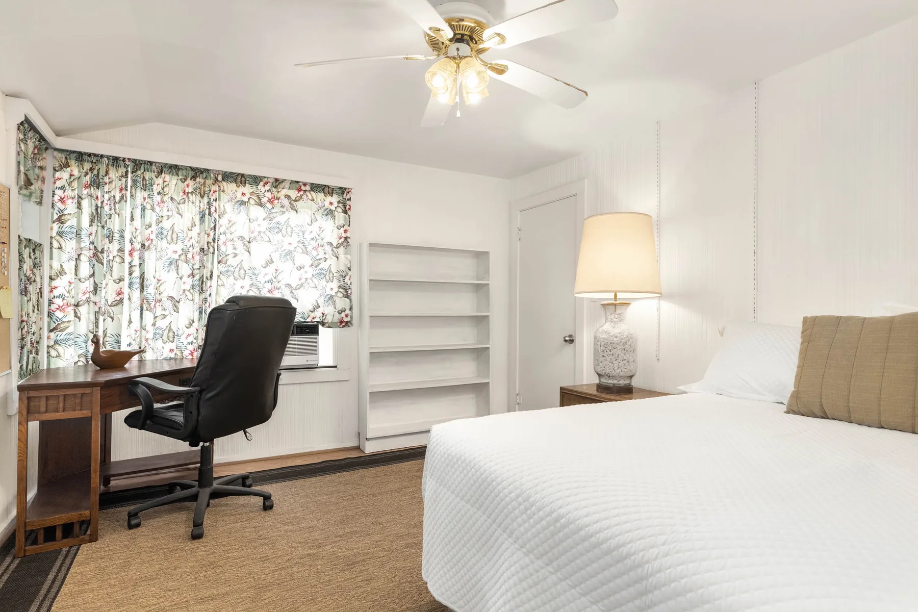 A bedroom with a desk and chair beside a window with floral curtains.