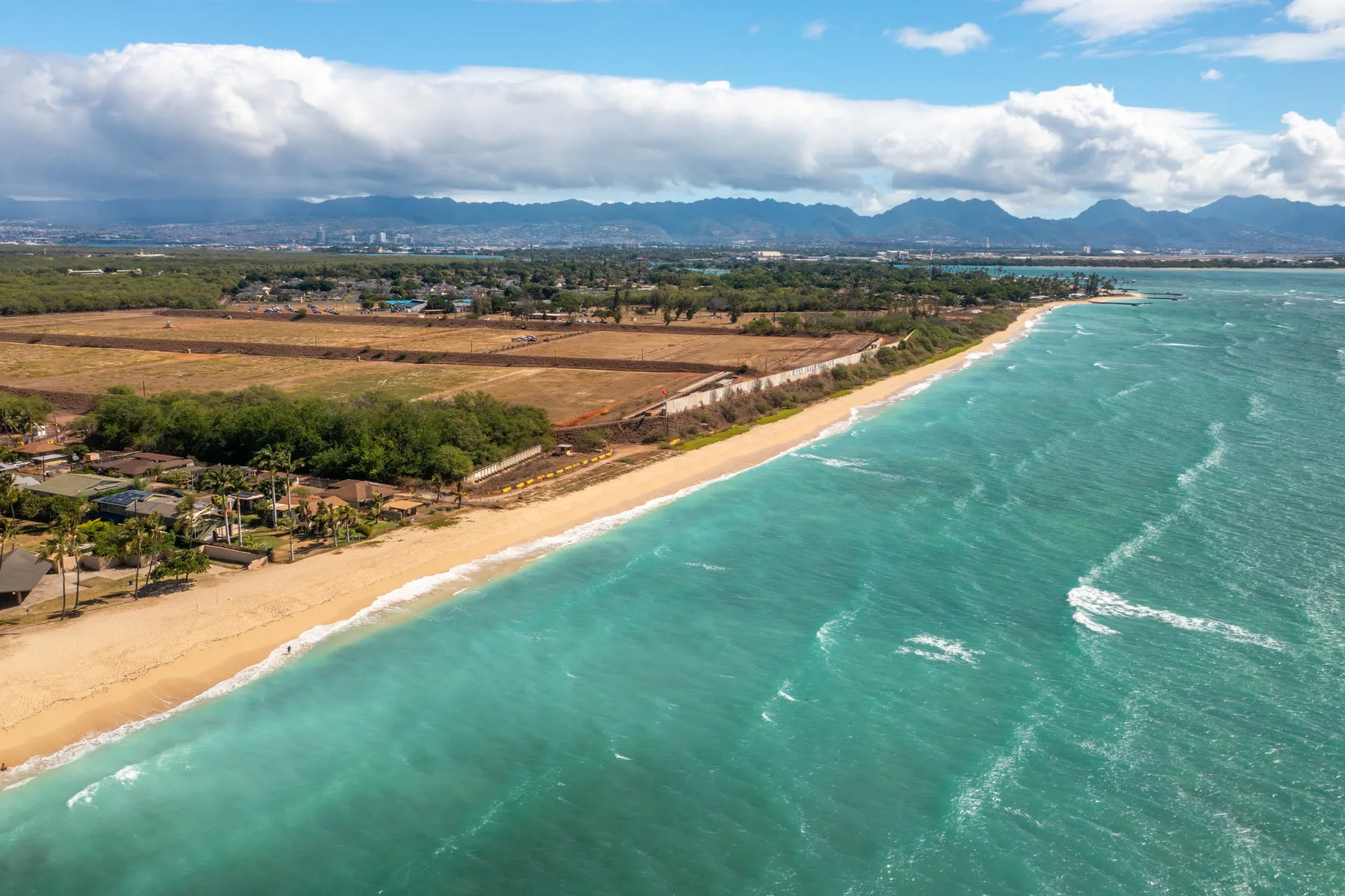 Aerial view of a sandy coastline with turquoise water and fields inland.
