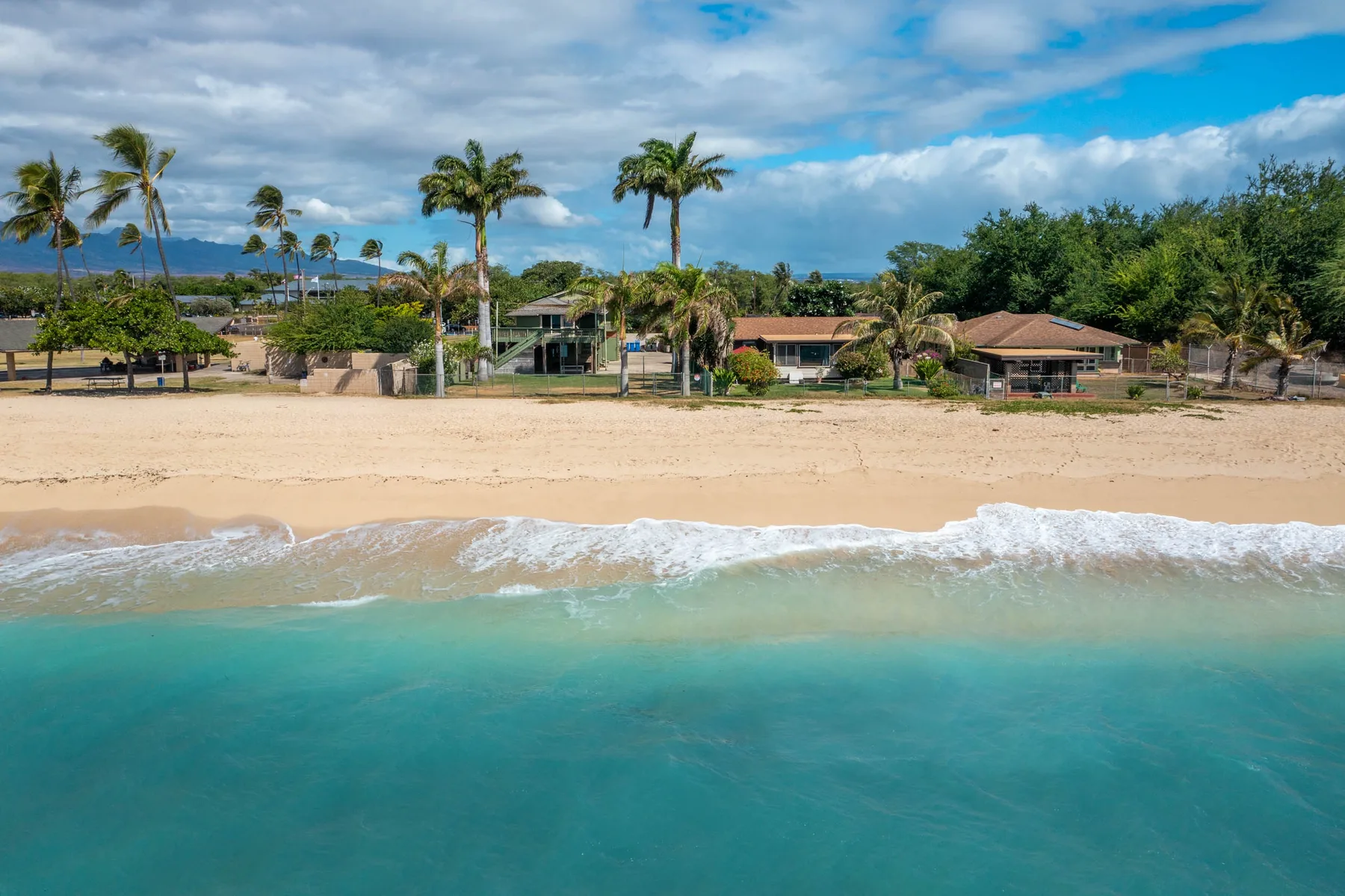 A sandy beach meets turquoise water, with palm trees and buildings along the shore.