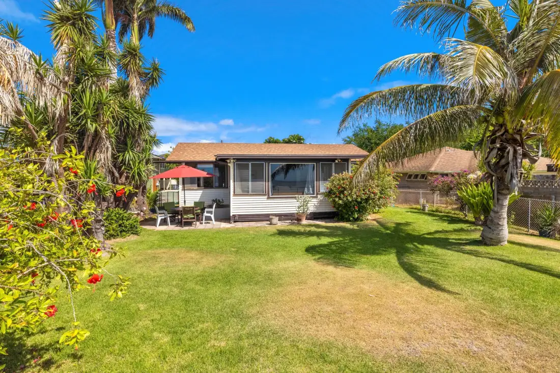 A small house sits beside a lawn, with palm trees and outdoor seating.