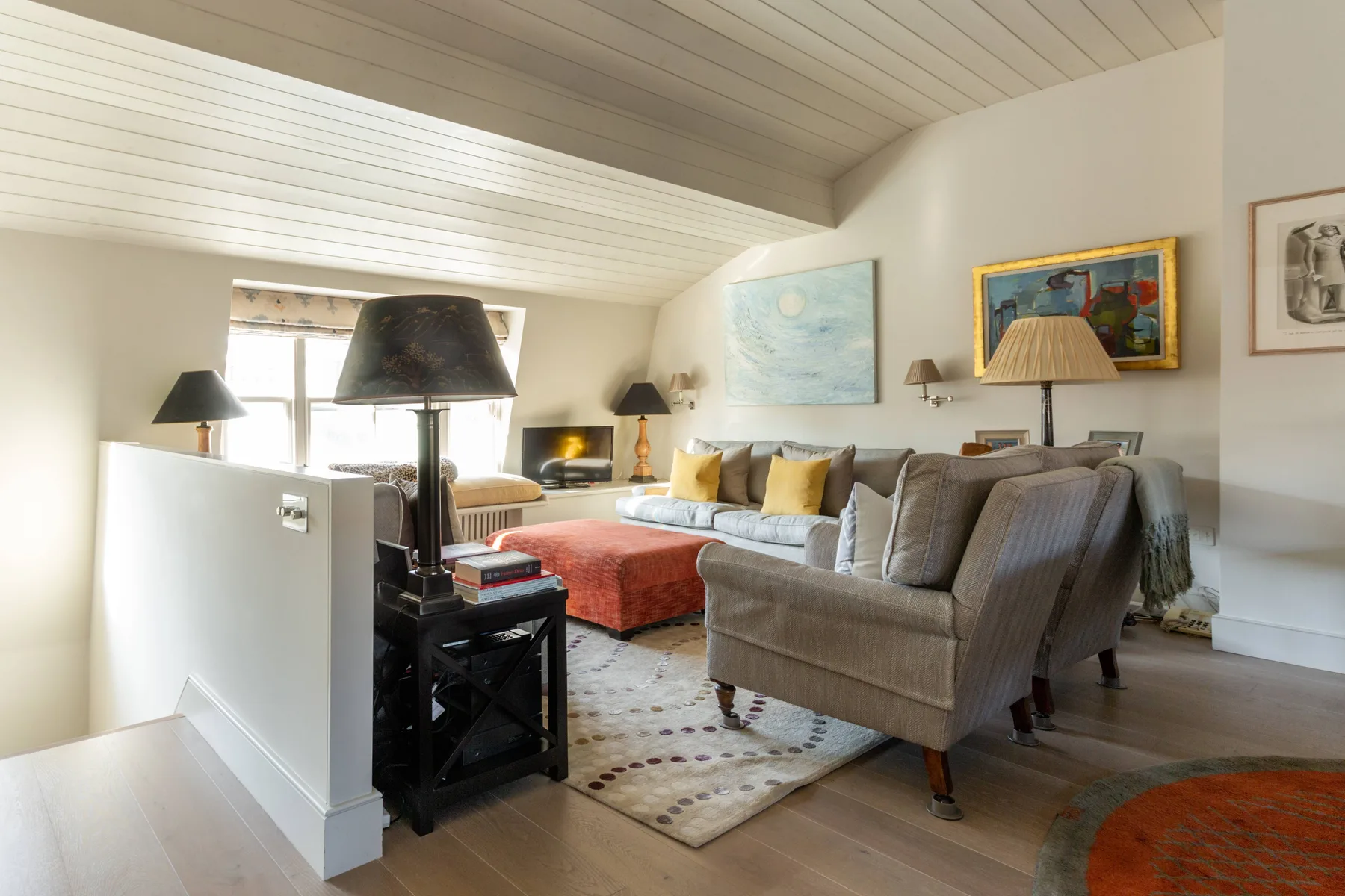 A bright living room with gray seating, a red ottoman, and colorful artwork.