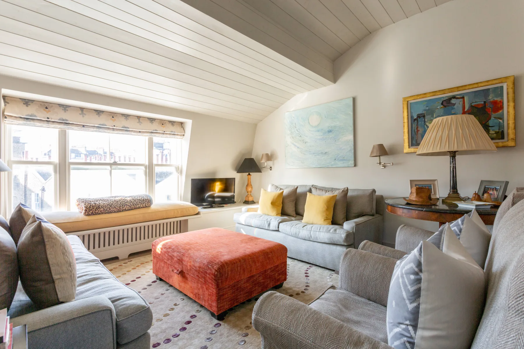 A bright living room with seating arranged around a red ottoman.