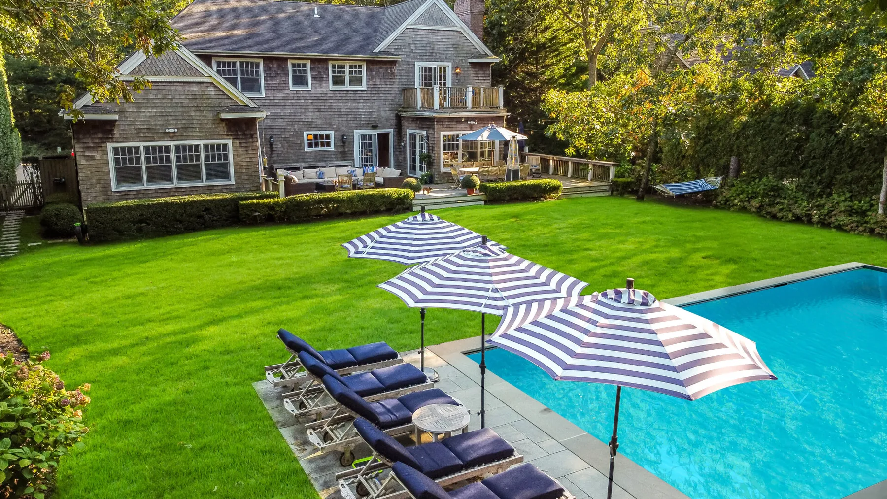 A landscaped backyard has a swimming pool, striped umbrellas, and lounge seating.