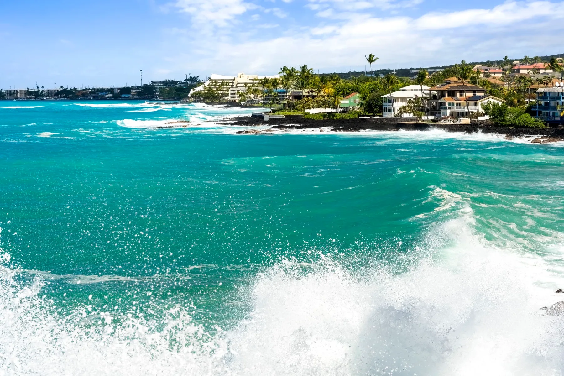Olas turquesas rompen junto a una costa tropical bordeada de edificios.