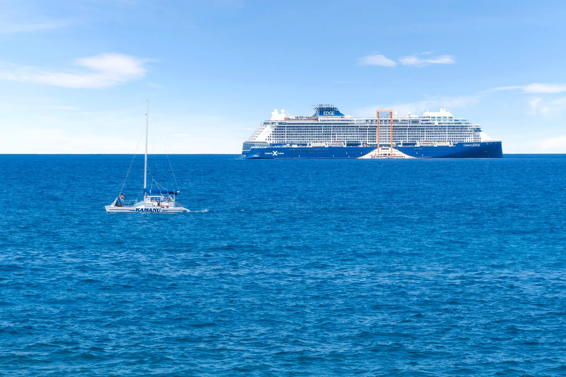 Un gran crucero y un pequeño velero se ven sobre el mar azul.