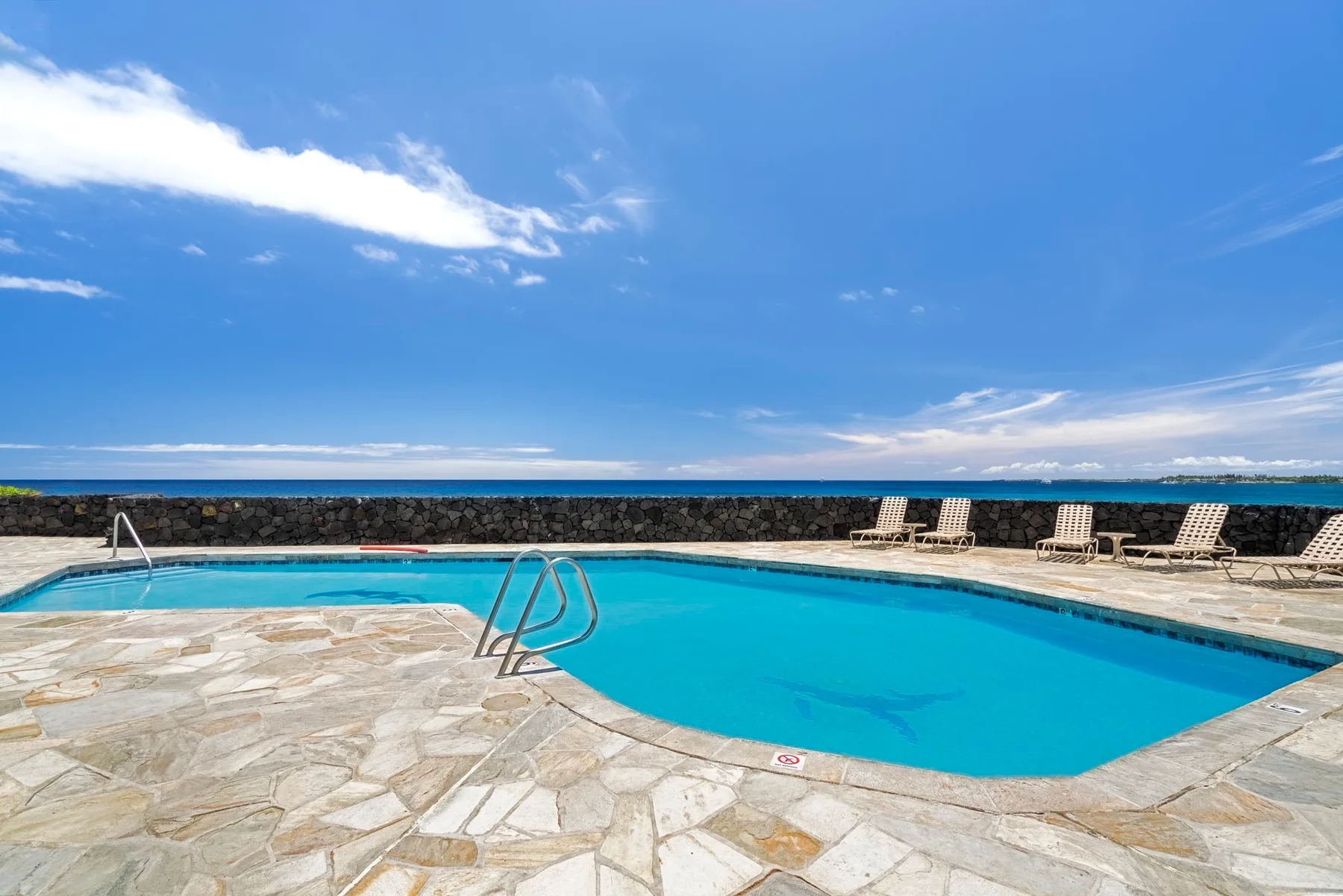 Una piscina al aire libre está junto a una terraza de piedra bajo un cielo azul.