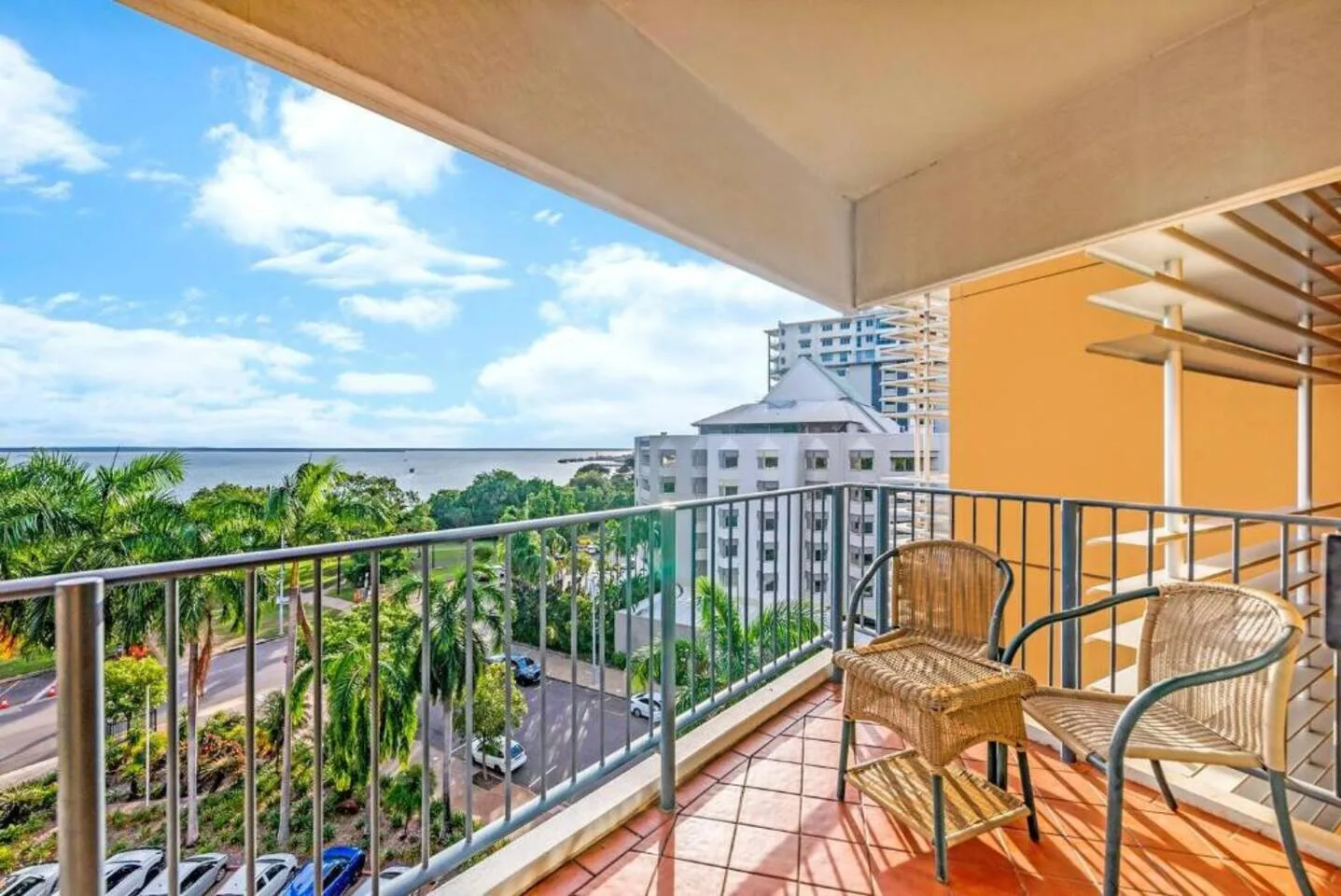 A covered balcony has outdoor seating overlooking palm trees and nearby buildings.