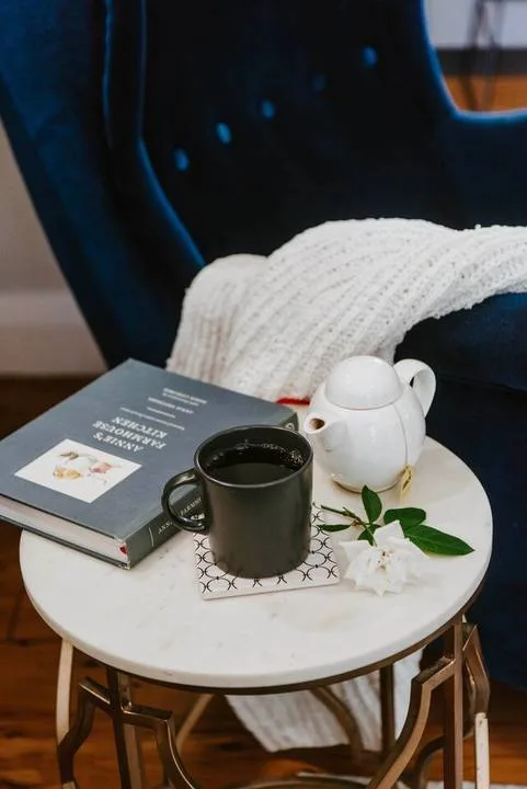 A small round table holds a mug, teapot, book, and flower beside a dark chair.