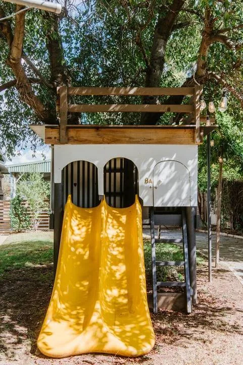 A wooden play structure with a bright yellow slide sits beneath leafy trees.