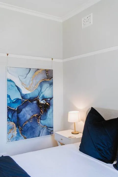 A bedroom with a bed, bedside table, lamp, and blue abstract wall art.