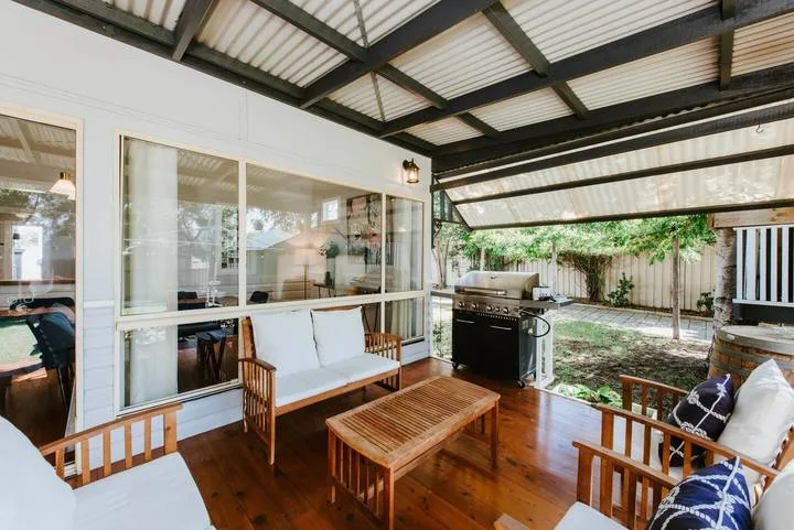 A covered outdoor patio has wooden seating and a barbecue beside the house.