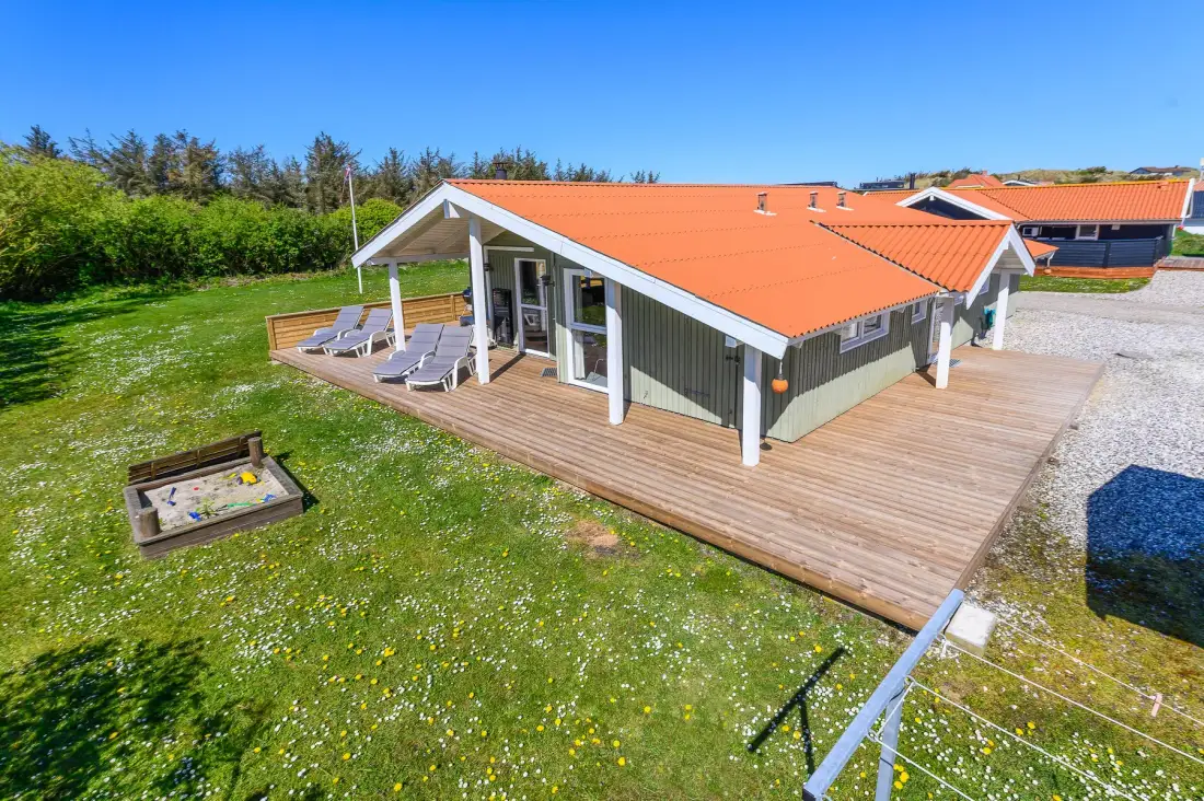 Una casa moderna con techo naranja junto a una amplia terraza de madera y un jardín con césped.