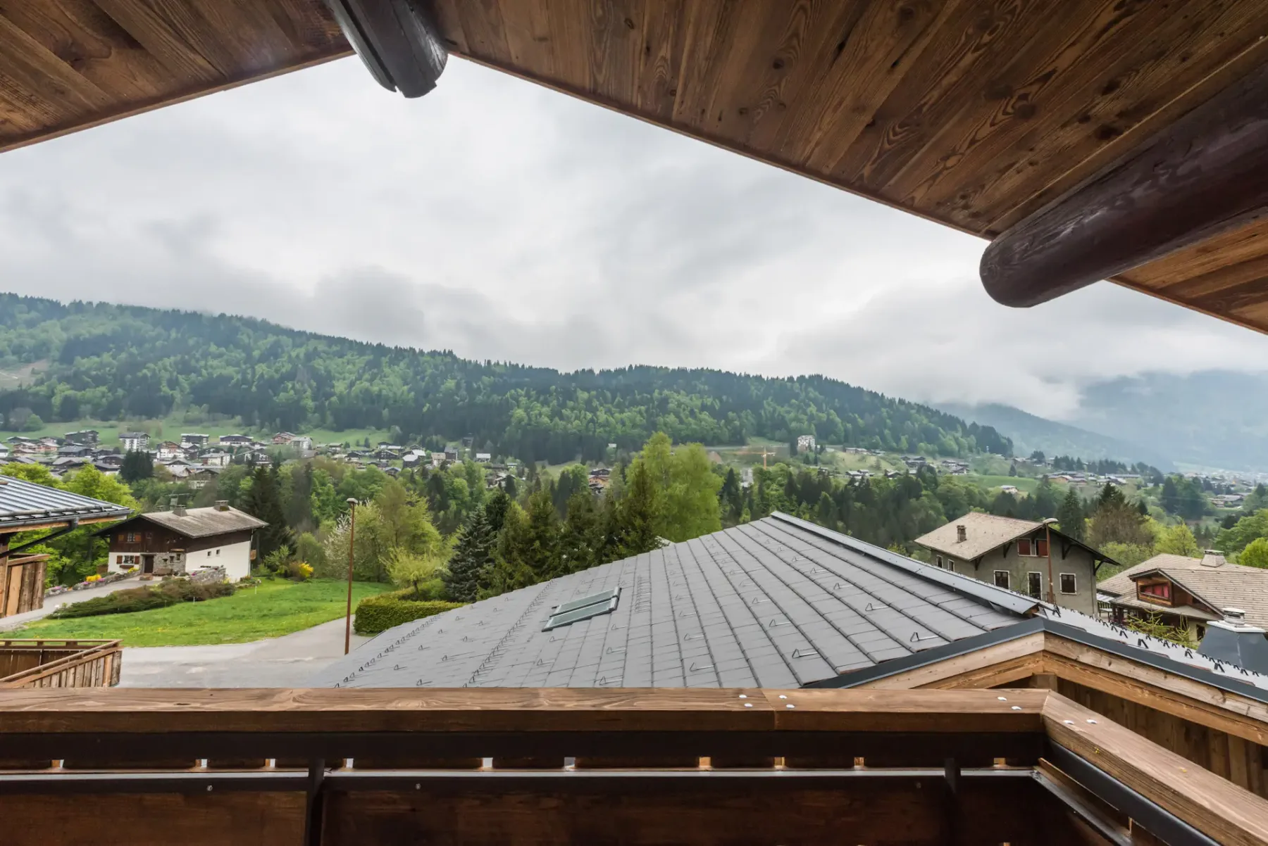 Una terraza de madera da a tejados, un pueblo y colinas verdes bajo un cielo nublado.