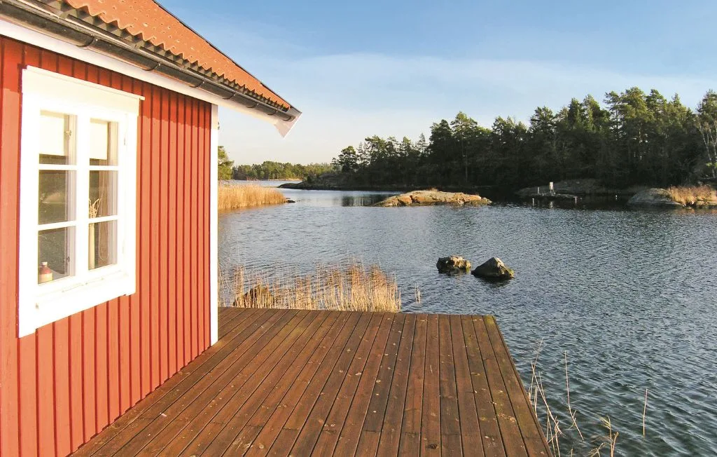 Una terraza de madera junto a un lago, con un edificio rojo y árboles en la orilla opuesta.