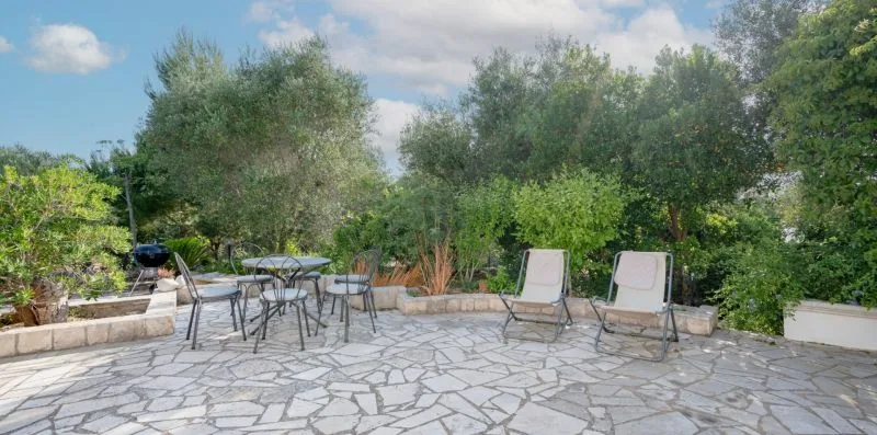 A stone-paved patio with outdoor seating, surrounded by leafy trees and plants.