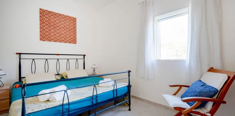 A bedroom with a metal-framed bed and a chair beside a bright window.